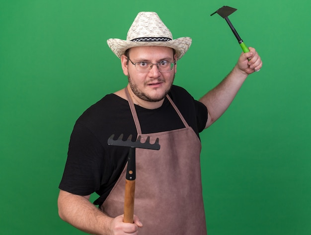 Pleased young male gardener wearing gardening hat holding rake with hoe rake standing in fighting pose isolated on green wall