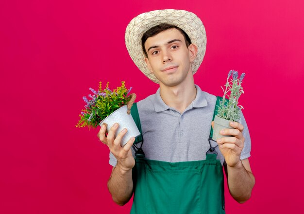 Pleased young male gardener wearing gardening hat holds flowers in flowerpots