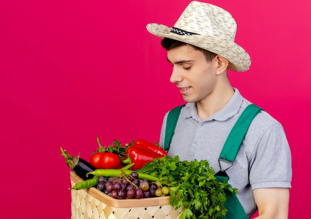 Pleased young male gardener wearing gardening hat holds and looks at vegetable basket 