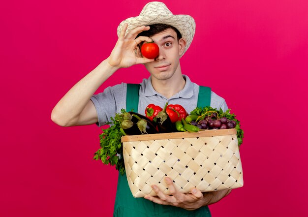 ガーデニング帽子をかぶって喜んでいる若い男性の庭師は野菜のバスケットを保持します