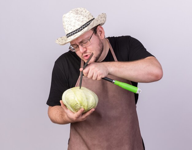 Pleased young male gardener wearing gardening hat scratching cabbage with hoe rake isolated on white wall