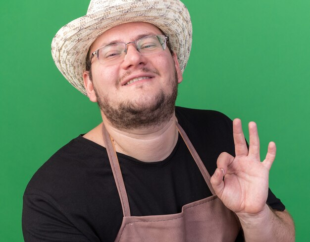 Free photo pleased young male gardener wearing gardening hat showing okay gesture isolated on green wall