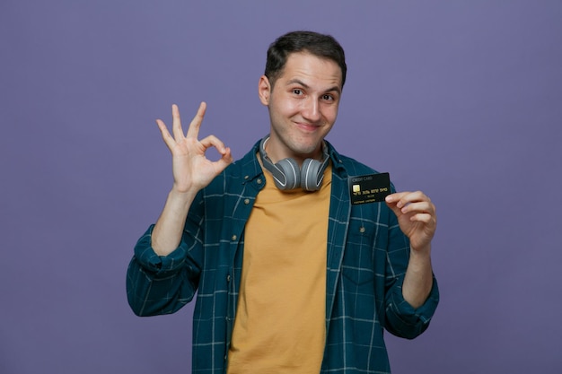 Pleased young male student wearing headphones around neck looking at camera showing credit card and ok sign isolated on purple background
