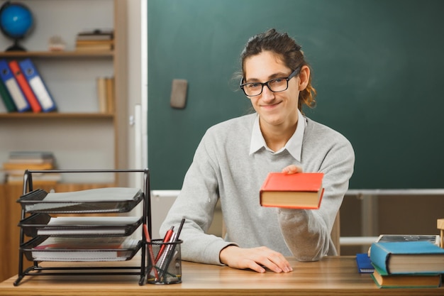 pleased young male teacher holding out book at camera sitting at desk with school tools on in classroom