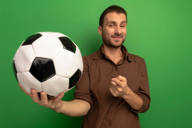 Felice giovane uomo allungando il pallone da calcio verso la parte anteriore guardando la telecamera puntata verso la palla isolata sulla parete verde