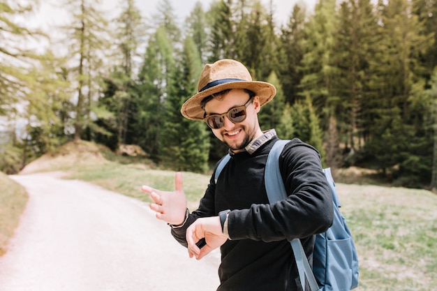 Felice giovane uomo in occhiali da sole guardando l'orologio da polso con il sorriso in piedi sulla strada sulla natura