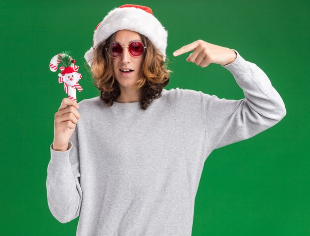 クリスマスのサンタの帽子と赤い眼鏡を身に着けている若い男は、緑の背景の上に立っているそれで人差し指を指して笑顔のクリスマスキャンディケインを持って喜んで