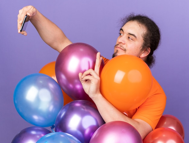 Pleased young man wearing orange t-shirt standing behind balloons take a selfie showing peace gesture isolated on purple wall