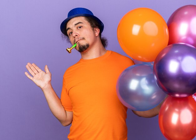 Pleased young man wearing party hat holding balloons blowing party whistle spreading hand isolated on purple wall