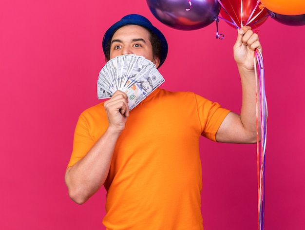 Free photo pleased young man wearing party hat holding balloons and covered face with cash