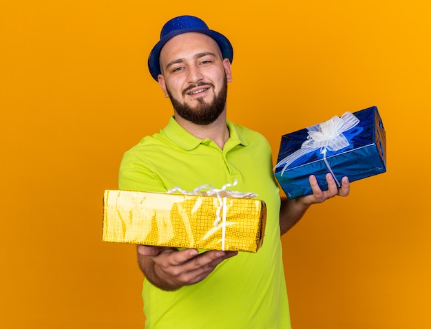 Pleased young man wearing party hat holding out gift boxes at camera 