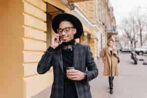 Бесплатное фото Довольный молодой человек с коричневой кожей пьет кофе на улице. счастливый африканский парень с чашкой латте, называя кого-то.