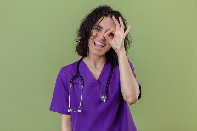 Pleased young nurse wearing uniform and stethoscope smiling cheerfully making ok sign looking through this sign standing on isolated green