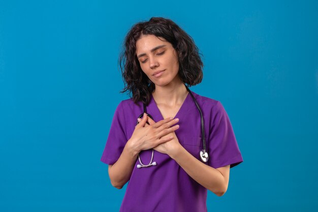 Довольная молодая медсестра в униформе и стетоскопе, стоя со скрещенными руками на груди, жестом ощущает положительные эмоции на изолированном синем