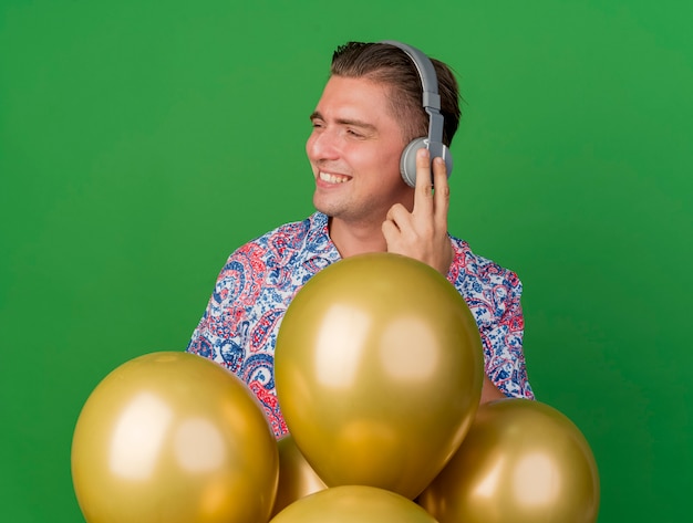 Foto gratuita piacere giovane partito ragazzo guardando a lato indossando una maglietta colorata e cuffie in piedi dietro palloncini isolati su verde