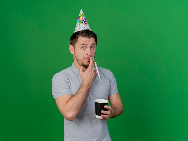 緑で隔離のコーヒーのカップを保持している笛を吹く誕生日のキャップを身に着けている若いパーティーの男を喜ばせる