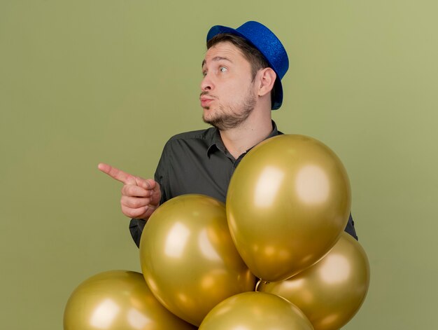 pleased young party guy wearing black shirt and blue hat standing behind balloons and points at side isolated on olive green