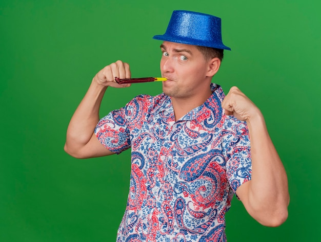 Бесплатное фото Довольный молодой тусовщик в синей шляпе дует вечеринку, показывая сильный жест, изолированный на зеленом