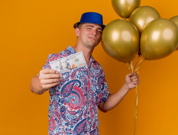 Free photo pleased young party guy wearing blue hat holding balloons and holding out cash isolated on orange