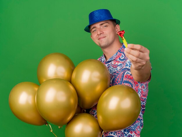 풍선을 들고 녹색에 고립 된 파티 송풍기를 들고 파란색 모자를 쓰고 기쁘게 젊은 파티 남자