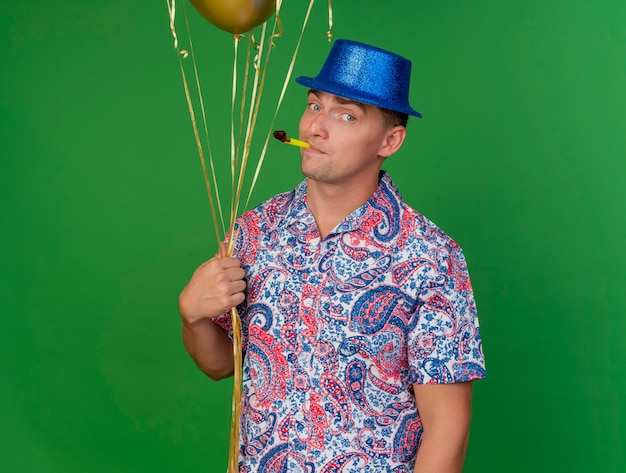 風船を保持し、緑で隔離の口にパーティーブロワーを入れて青い帽子をかぶって喜んで若いパーティー男