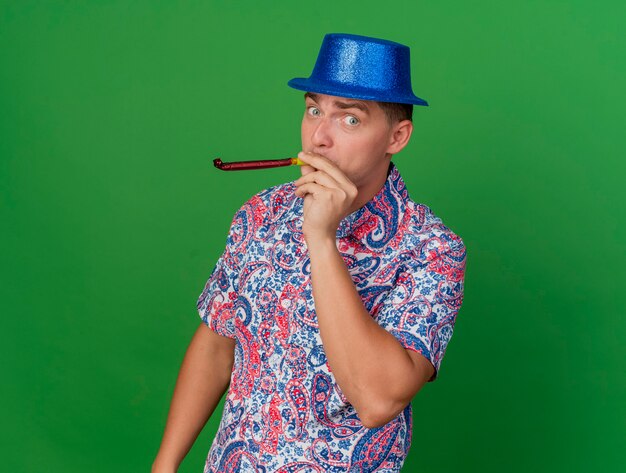파란색 모자를 입고 녹색에 고립 된 파티 송풍기를 불고 기쁘게 젊은 파티 남자