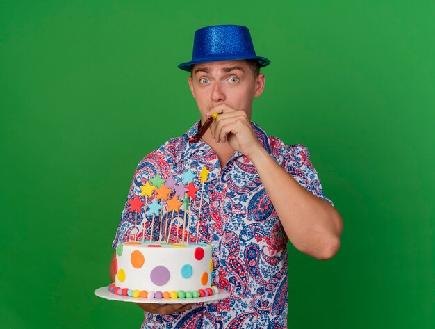 ケーキを保持し、緑に分離されたパーティーブロワーを吹く青い帽子をかぶって喜んで若いパーティー