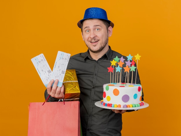 Pleased young party guy wearing blue hat holding cake with gifts and tickets isolated on orange