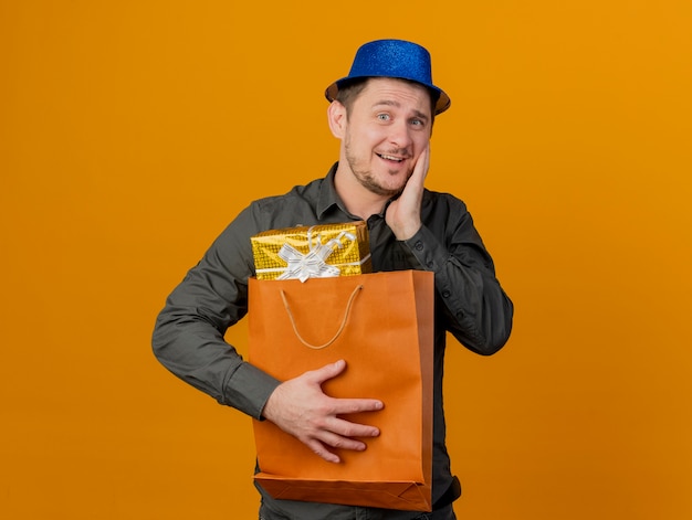 Free photo pleased young party guy wearing blue hat holding gift bag and putting hand on cheek isolated on orange