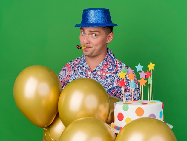녹색에 고립 된 파티 송풍기를 불고 케이크를 들고 풍선 뒤에 서있는 파란색 모자를 쓰고 기쁘게 젊은 파티 남자