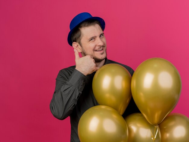 Pleased young party guy wearing blue hat standing next to balloons showing phone call gesture isolated on pink