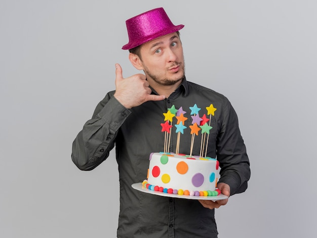 흰색에 고립 된 전화 제스처를 보여주는 케이크를 들고 분홍색 모자를 쓰고 기쁘게 젊은 파티 남자