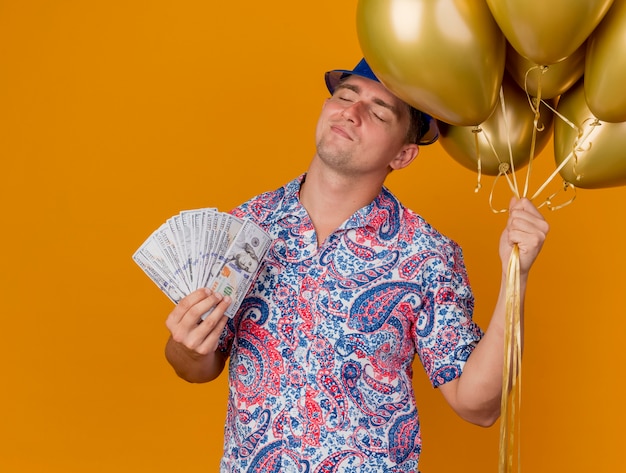 Pleased young party guy with closed eyes wearing blue hat holding balloons and cash isolated on orange