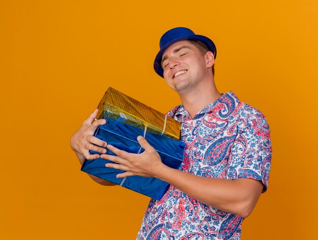 Pleased young party guy with closed eyes wearing blue hat hugged gift boxes isolated on orange