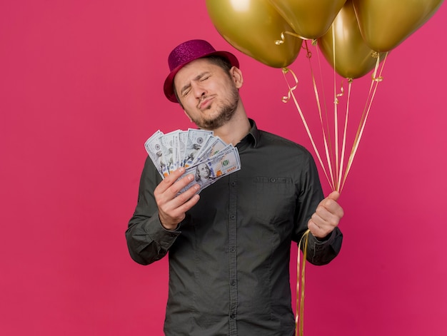 Довольный молодой тусовщик с закрытыми глазами в розовой шляпе держит деньги с воздушными шарами, изолированными на розовом
