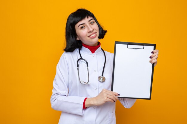 Pleased young pretty caucasian girl in doctor uniform with stethoscope holding clipboard and 