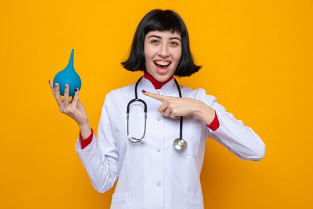 Pleased young pretty caucasian girl in doctor uniform with stethoscope holding and pointing at enema