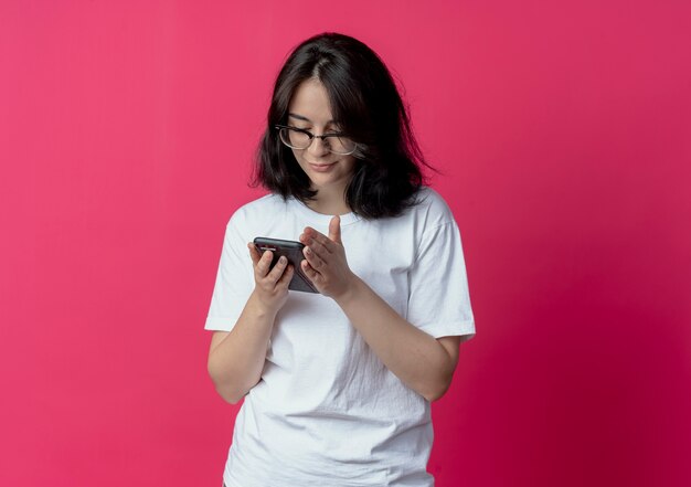 Довольная молодая красивая кавказская девушка держит и смотрит на мобильный телефон, изолированные на малиновом фоне с копией пространства