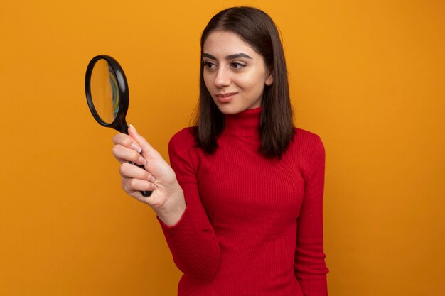 Pleased young pretty caucasian girl holding magnifying glass looking at side through magnifying glass 