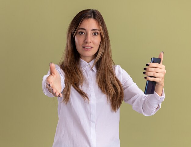 Pleased young pretty caucasian girl holding phone and stretching out hand on olive green 