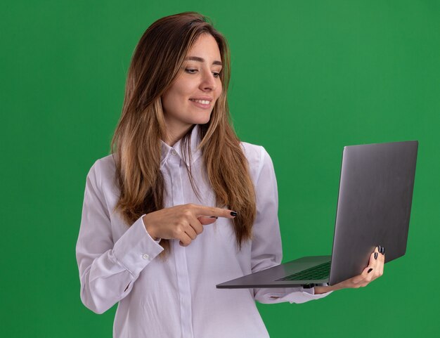 Pleased young pretty caucasian girl looks and points at laptop isolated on green wall with copy space