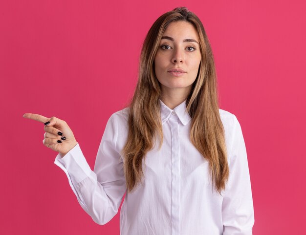 Pleased young pretty caucasian girl points at side on pink 