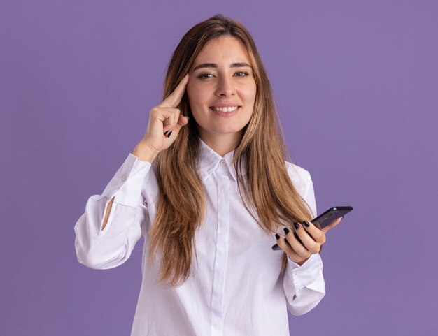 Pleased young pretty caucasian girl puts finger on temple and holds phone