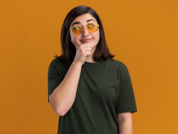 Pleased young pretty caucasian girl in sun glasses holds chin and looks up on orange