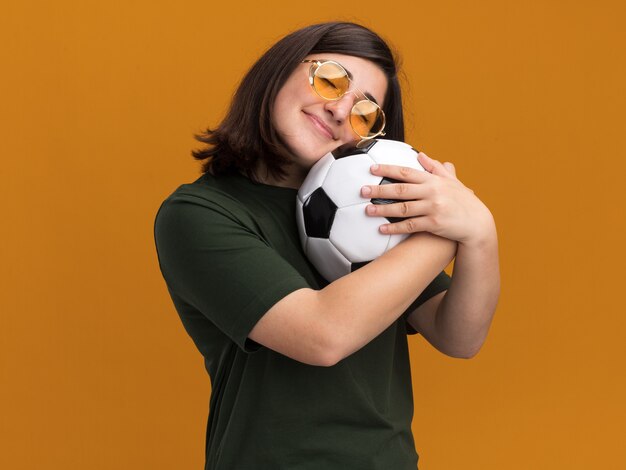 Pleased young pretty caucasian girl in sun glasses hugs ball isolated on orange wall with copy space