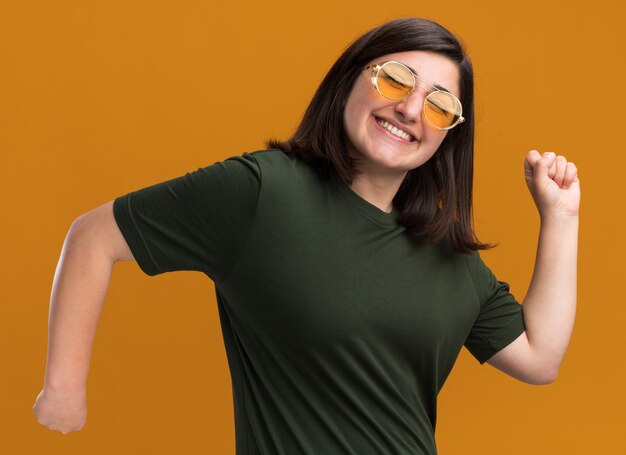 Pleased young pretty caucasian girl in sun glasses stands sideways keeping fists isolated on orange wall with copy space