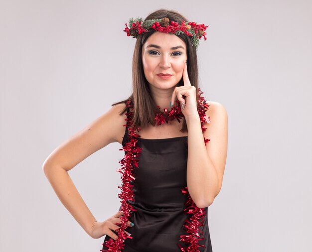 Pleased young pretty caucasian girl wearing christmas head wreath and tinsel garland around neck looking at camera keeping hand on waist touching face isolated on white background with copy space