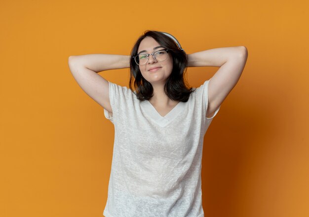 Pleased young pretty caucasian girl wearing glasses and headphones putting hands behind head isolated on orange background with copy space