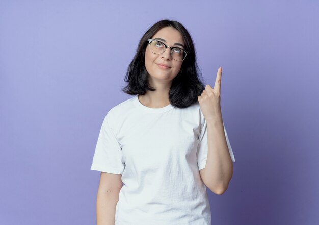 Pleased young pretty caucasian girl wearing glasses looking and pointing up isolated on purple background with copy space