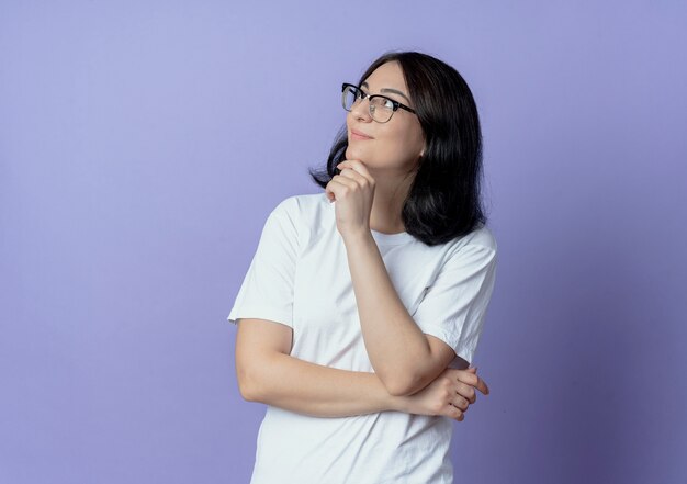 Pleased young pretty caucasian girl wearing glasses putting hands under elbow and on chin looking at side
